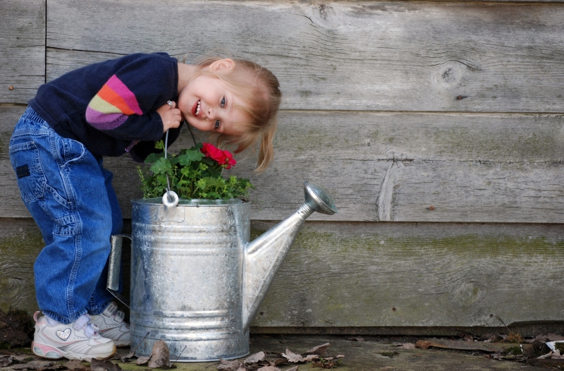 En glad flicka står lutad över och håller i en vattenkanna i zink. I vattenkannan är det planterade växter