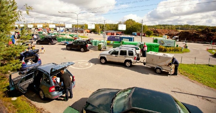 Bilar står parkerade vid en återvinningscentral. Människor tömmer sina bilar på saker som kan återvinnas.