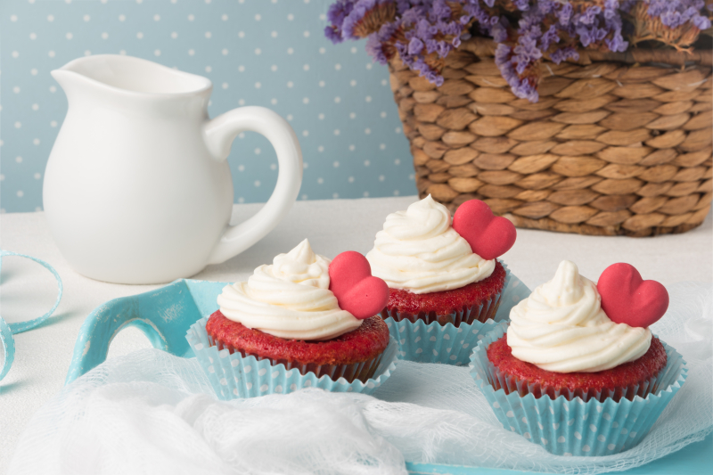 Tre cupcakes med hjärtan på är framdukade på ett bord. På bordet står också en flätad blomkruka med lila blommor i och en vit mjölkkanna.