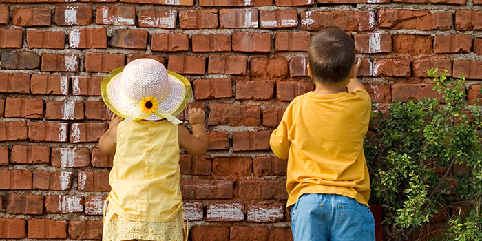 En pojke och en flicka står mot en röd tegelvägg. Flickan har en vit sommarhatt på sig. Båda barnen har gula kläder