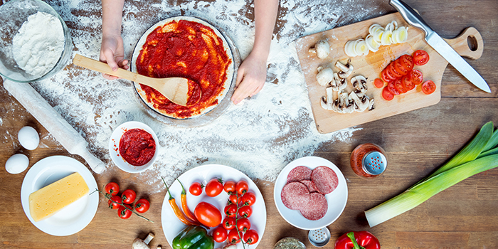 Ett barn gör en hemmagjord pizza på ett träbord. På bordet står ingredienser framdukade. Det är salamiskivor, purjolök, tomatskivor, champinjonskivor, paprika, ost och tomatsås.