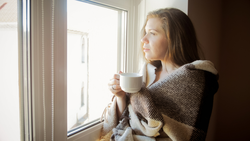 En kvinna står och tittar ut genom ett fönster. I handen har hon en vit kaffekopp. Hon har en filt runt axlarna.