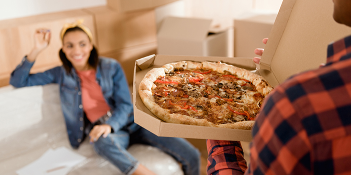 En leende kvinna sitter på en soffa och tittar på en man som håller i en pizzakartong. I bakgrunden står det flera flyttkartonger.