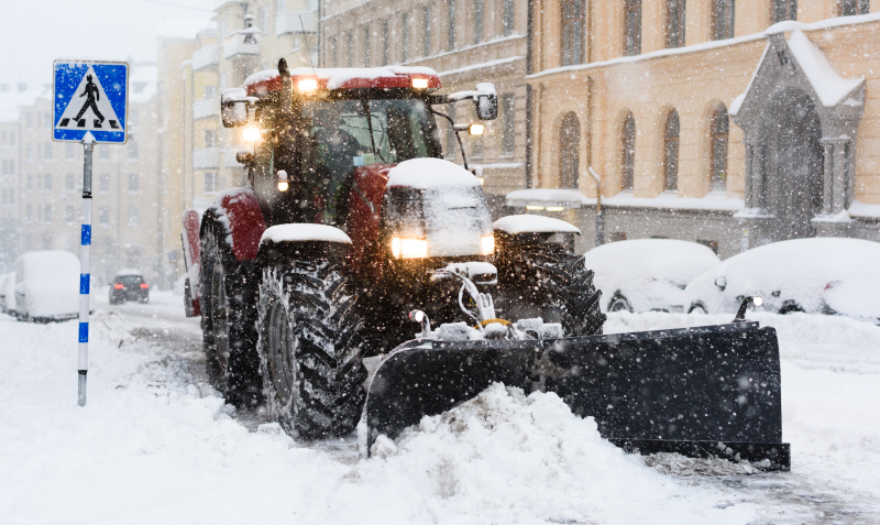 En röd snöröjare plogar en stadsgata täckt av snö. Längs gatan står det insnöade bilar på rad