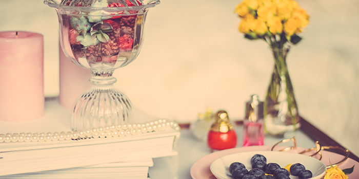 Vintage heminredning med en glasskål med något ätbart i, ett pärlhalsband ligger framför skålen ovanpå två böcker. En vas med gula rosor står i bakgrunden och ett fat med blåbär omges av två små parfymflaskor och ett guldarmband.