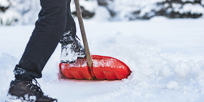 Marken är fylld av snö. Nederdelen av en människas ben och ett par snöiga kängor syns. Den ena foten är ovanpå en röd snöskyffel.