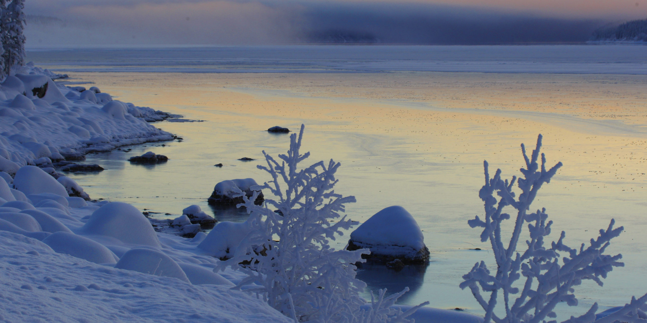 Nysnö täcker marken, stenarna och trädens grenar. Intill land, på och runt stenar som är svarta i motljuset, ligger isen pärlemorskimrande. Längre ut ligger dimman som solen färgar i regnbågens alla färger.