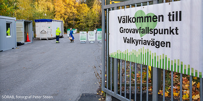 En skylt med välkommen till Grovafallspunkt Valkyriavägen sitter på entrégrinden. Två människor samtalar om avfall framför avfallscontainrar.