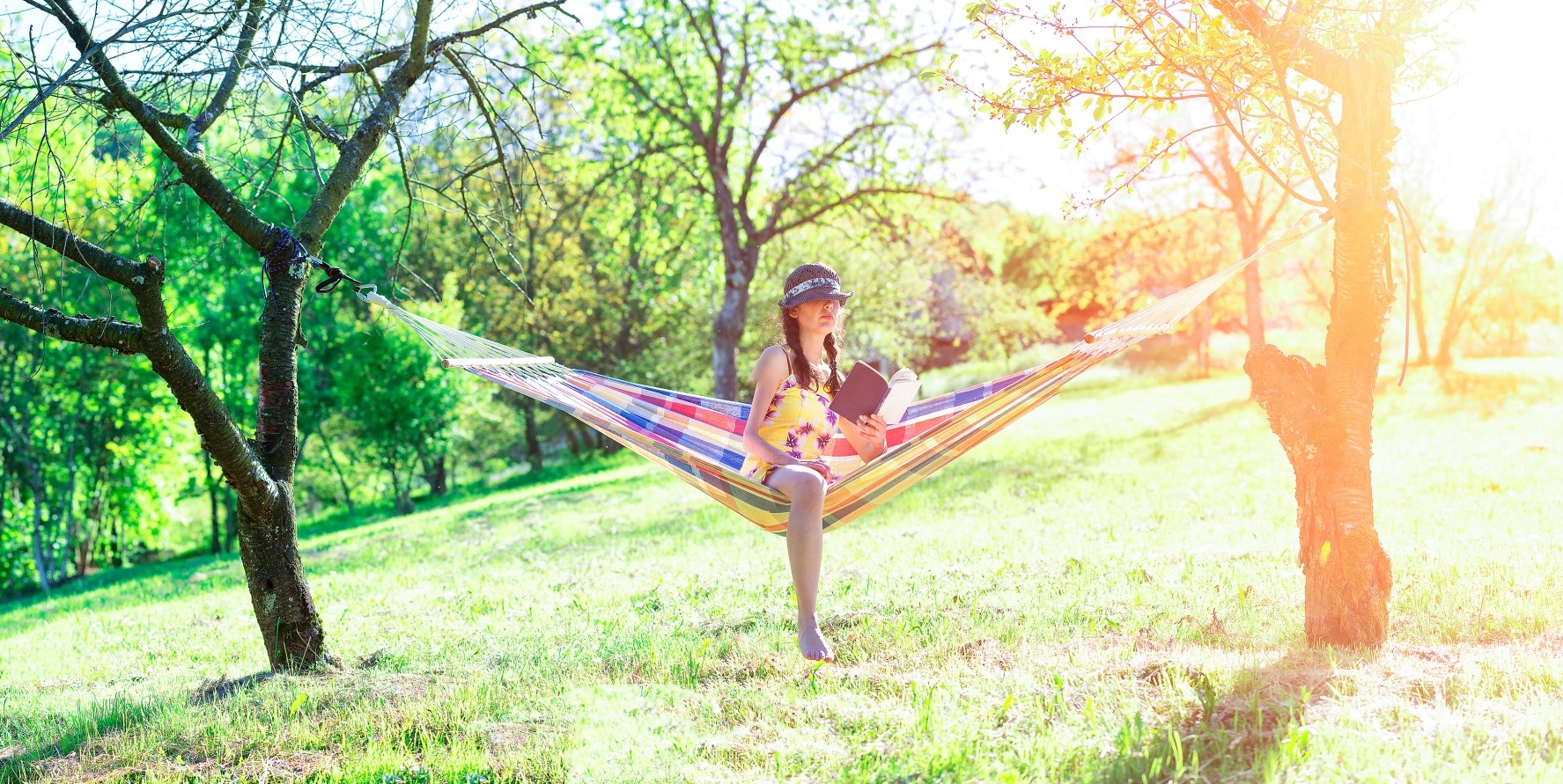En kvinna sitter i en hängmatta mellan två träd. I handen håller hon i en bok. Hon har en hatt på huvudet. I bakgrunden skiner solen.