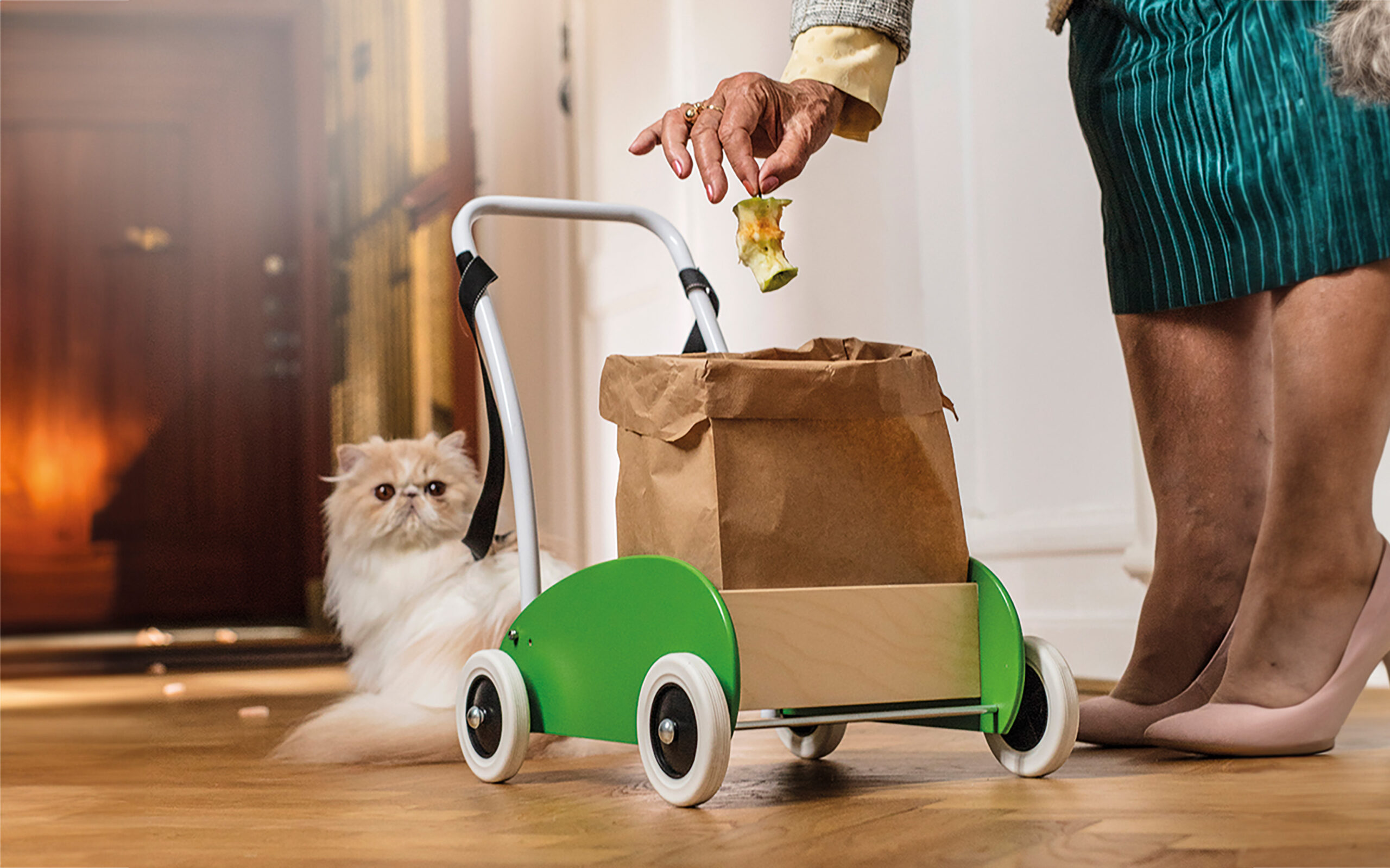 En dam med kjol och högklackade skor lägger ner ett äppleskrott i en matavfallspåse som står på en "lära-gå-vagn". Bakom vagnen ligger en vit/beige katt.