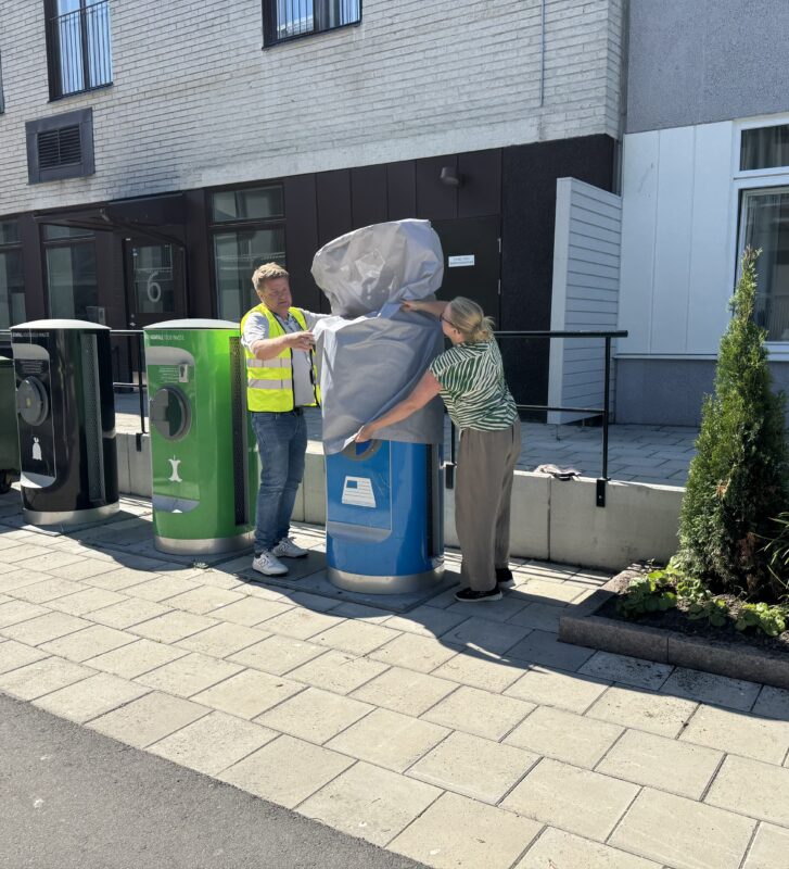 Två personer som trär en grå plasthuva över ett sopsugsinkast en solig dag, Inkasten är blåa och gröna